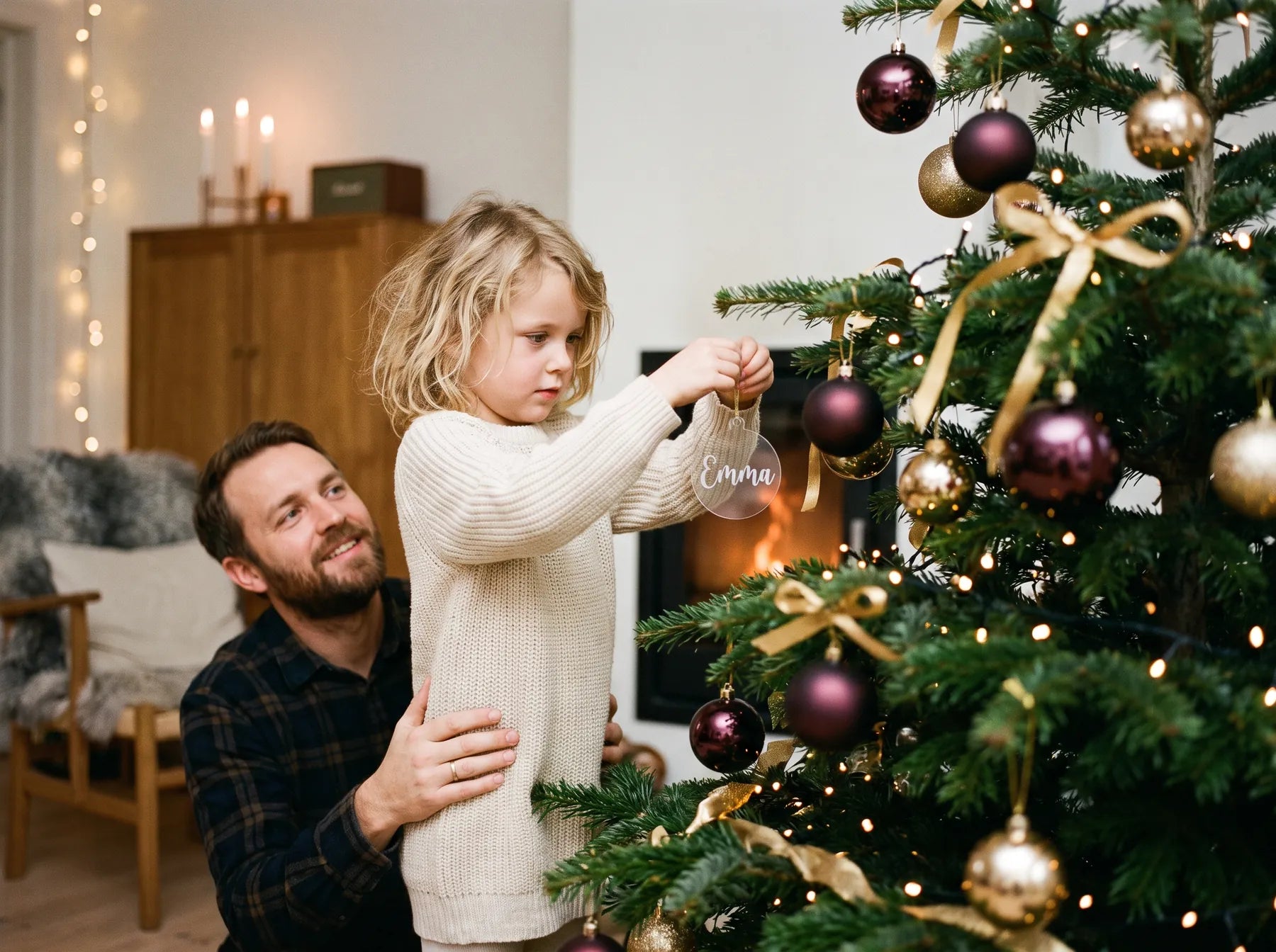 En pige hænger en julekugle i klar akryl med navnet Emma på juletræet, mens hendes far holder om hende